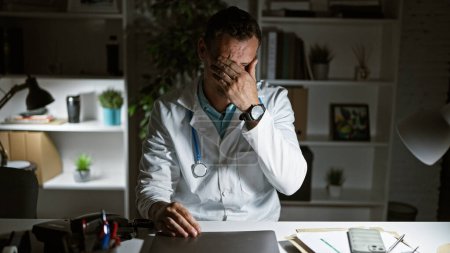 Ein gestresster Mann in weißem Laborkittel sitzt an einem Schreibtisch in einem Krankenhauszimmer und schildert Erschöpfung oder Besorgnis.