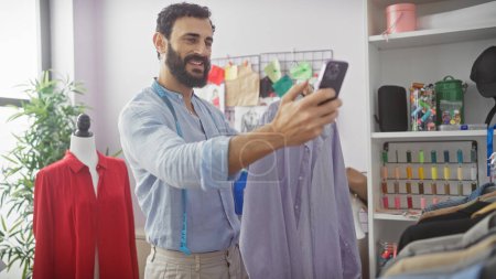 Foto de Sonriente hombre barbudo tomando selfie en sastrería rodeado de prendas de vestir e hilos de colores - Imagen libre de derechos