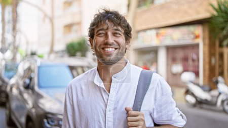 Foto de Un hombre hispano sonriente con ojos verdes y barba, parado al aire libre en un entorno urbano soleado con una camisa blanca y llevando una bolsa. - Imagen libre de derechos