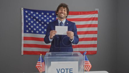 Smiling hispanic man with a beard holding a ballot at american electoral voting station with us flags.