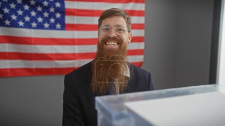 Smiling bearded man with glasses in a suit against american flag backdrop in a well-lit office