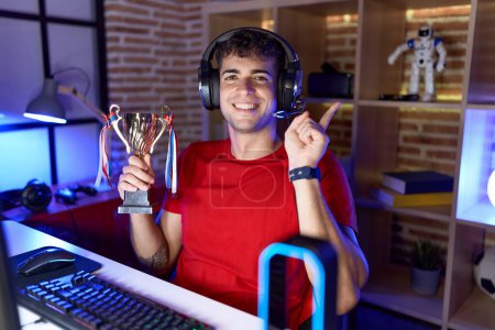 Foto de Joven hispano jugando videojuegos sosteniendo trofeo sonriendo feliz apuntando con la mano y el dedo hacia un lado - Imagen libre de derechos