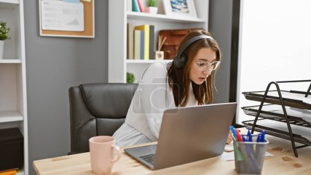 Photo for A focused young woman works attentively in a modern office, wearing headphones and eyeglasses, with a laptop, mug, and stationery on her desk. - Royalty Free Image