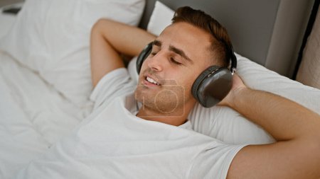 Photo for Relaxed hispanic man enjoying music with headphones in a cozy bedroom setting - Royalty Free Image