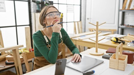 Une menuisière réfléchie avec des lunettes de sécurité dans un atelier de menuiserie entouré d'outils et d'objets en bois.