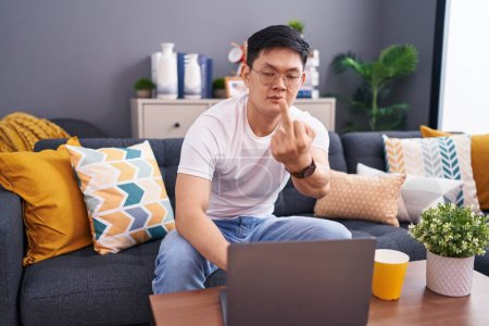 Foto de Young asian man using laptop at home sitting on the sofa showing middle finger, impolite and rude fuck off expression - Imagen libre de derechos