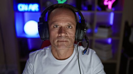 Seriöser Mann mittleren Alters, der in seinem Home Office in die digitale Welt des Spielens eintaucht - gutaussehender hispanischer Gamer, rockige Kopfhörer, die ganze Nacht Cyber-Unterhaltung streamen