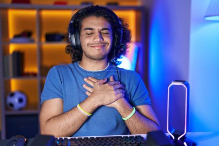 Hispanischer Mann mit lockigem Haar, der Videospiele spielt, mit den Händen auf der Brust lächelnd, die Augen geschlossen mit dankbarer Geste im Gesicht. Gesundheitskonzept. 