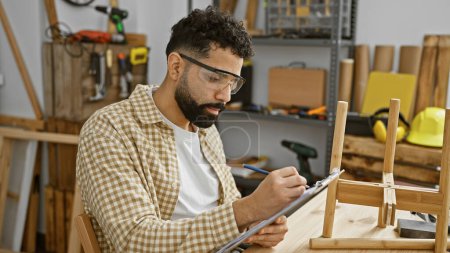 Handsome hispanic carpenter with beard takes notes in a well-equipped woodworking workshop.