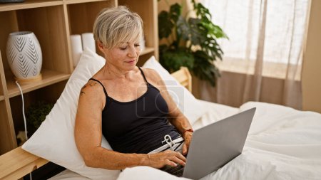Téléchargez les photos : Femme mûre aux cheveux gris utilisant un ordinateur portable dans un cadre confortable de chambre à coucher à la maison, rayonnant de confort et de productivité. - en image libre de droit