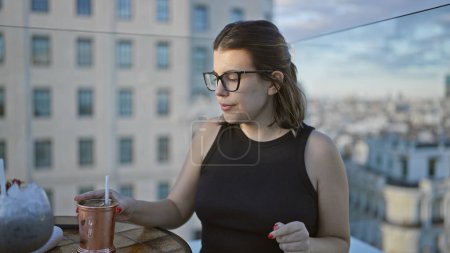 Beautiful hispanic woman, donning glasses, savors a tropical cocktail on a rooftop terrace, a bubbly celebration high above the city