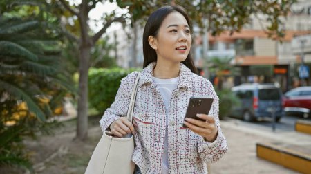Téléchargez les photos : Jeune femme asiatique en utilisant smartphone sur la rue de la ville, entouré de verdure et de véhicules. - en image libre de droit