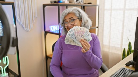 A mature woman in a purple sweater holds polish zloty in a well-lit gaming room, smiling behind the cash.