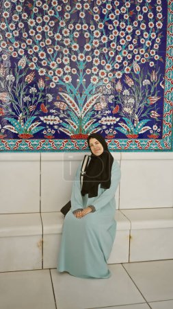 Una joven sonriente en un hiyab sentada frente a un decorado fondo floral islámico en una mezquita en los emiratos.