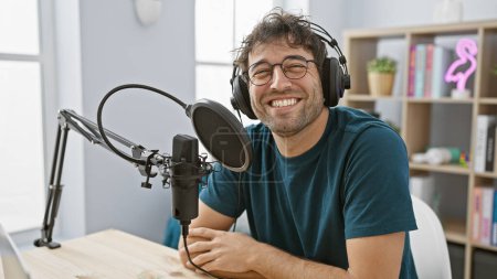 Téléchargez les photos : Homme hispanique avec écouteurs souriant en configuration studio radio avec microphone et bouclier pop - en image libre de droit