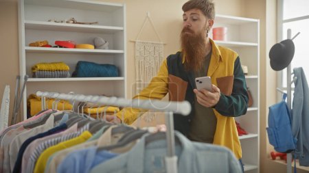 Photo for Redhead bearded man using smartphone in a modern tailor shop with colorful clothing - Royalty Free Image