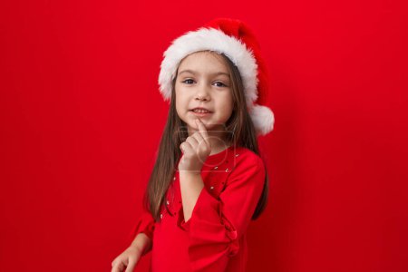Photo for Adorable hispanic girl in christmas hat, looking at camera, smiling with confidence, arms crossed, hand on chin over red isolated background thinking positive ideas, posing proud and cheerful - Royalty Free Image