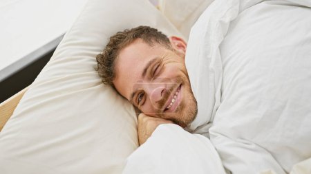Photo for A handsome adult man with a beard smiling while lying comfortably in white bed sheets in a home bedroom setting. - Royalty Free Image