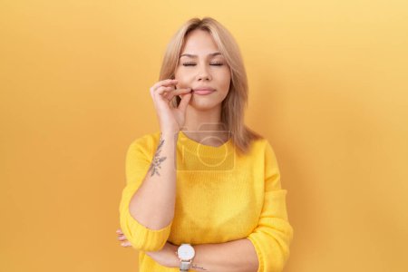 Jeune femme caucasienne portant un pull jaune bouche et lèvres fermées comme zip avec les doigts. secret et silencieux, tabou parlant 