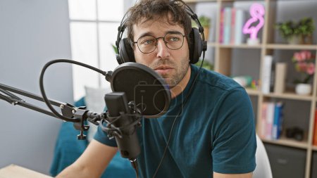 Ein junger bärtiger Mann mit Kopfhörern spricht in einem modernen Radiostudio in ein Mikrofon.