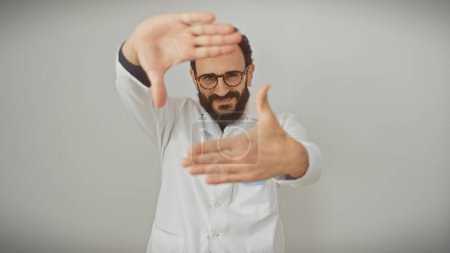 Photo for Smiling bearded man in white lab coat making frame gesture against isolated white background. - Royalty Free Image