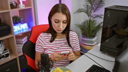 Foto de Una joven enfocada usa su teléfono inteligente en una vibrante sala de juegos con equipos modernos e iluminación colorida. - Imagen libre de derechos