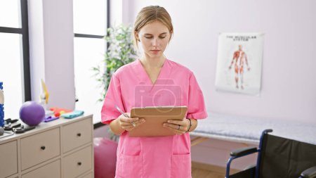 Foto de Una mujer caucásica enfocada usando un exfoliante médico rosa sosteniendo un portapapeles en una habitación clínica de rehabilitación brillante. - Imagen libre de derechos