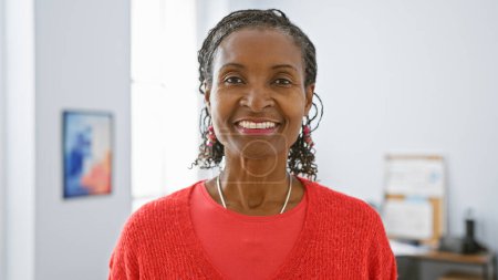 Téléchargez les photos : Femme africaine souriante en pull rouge posant avec confiance dans son bureau moderne, rayonnant de professionnalisme et de chaleur. - en image libre de droit