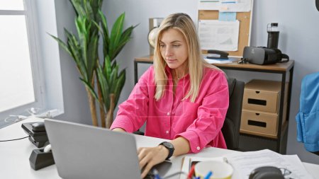 Foto de Una mujer rubia enfocada trabaja en un entorno de oficina, rodeada de comodidades modernas, tecnología y desorden de escritorio. - Imagen libre de derechos
