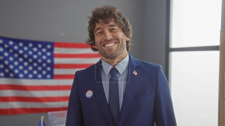 Joven hombre hispano sonriendo en el interior con la bandera americana de fondo vistiendo un traje y 'voté' pegatina