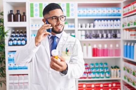 Foto de Joven farmacéutico latino sosteniendo pastillas botella hablando en el teléfono inteligente en la farmacia - Imagen libre de derechos