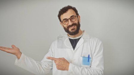 Smiling bearded man in white lab coat pointing aside, against a gray background, portrays professional advice or presentation.