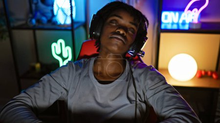 A serene young black woman enjoys music with headphones in a vibrant gaming room at night.
