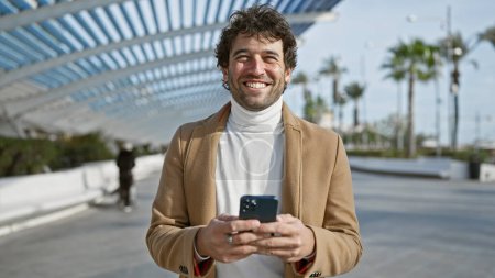 Ein hübscher junger hispanischer Mann lächelt, während er sein Smartphone im Freien in einem sonnigen, von Palmen gesäumten Stadtpark benutzt.