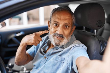 Téléchargez les photos : Homme âgé joyeux fait un selfie joyeux, pointant avec confiance et souriant avec la main et le doigt à sa voiture dans une rue urbaine - en image libre de droit