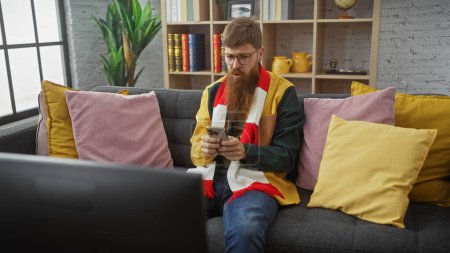Foto de Un joven barbudo usando intensamente un teléfono inteligente mientras descansa en un sofá en el interior rodeado de coloridas almohadas. - Imagen libre de derechos