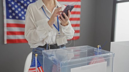 A young woman with a lanyard checks her cellphone in a voting center with a us flag and ballot box.