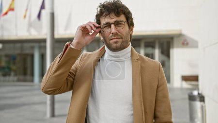 Photo for A stylish man adjusting glasses on an urban city street exudes confidence and fashion. - Royalty Free Image