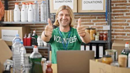 Smiling man with blond hair wearing a volunteer t-shirt gives thumbs up in a warehouse full of donations.
