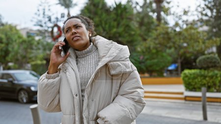 Foto de Una joven preocupada habla por teléfono mientras está parada en una calle de la ciudad, vistiendo un abrigo de invierno y rodeada de follaje urbano. - Imagen libre de derechos