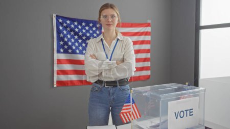 Téléchargez les photos : Jeune femme caucasienne avec les bras croisés se tient avec confiance dans une salle de collège nous électorale avec une urne et un drapeau. - en image libre de droit
