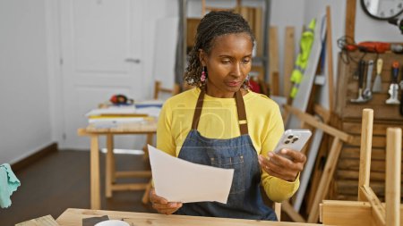 Photo for An african american woman reads a document and uses a smartphone in a carpentry workshop. - Royalty Free Image