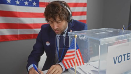 Téléchargez les photos : Un jeune homme concentré avec un casque marquant un bulletin de vote dans un bureau de vote américain, avec le drapeau US en arrière-plan. - en image libre de droit