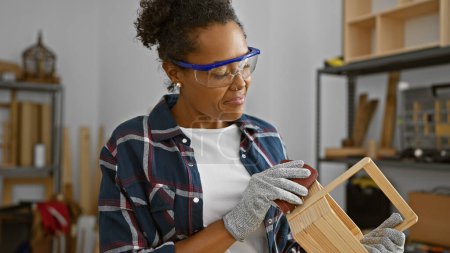 Photo for Focused woman sanding wood in a carpentry workshop, wearing safety glasses and gloves. - Royalty Free Image