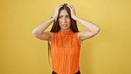 Photo for A perplexed young woman in an orange top clutching her head against a solid yellow background, expressing confusion and stress. - Royalty Free Image