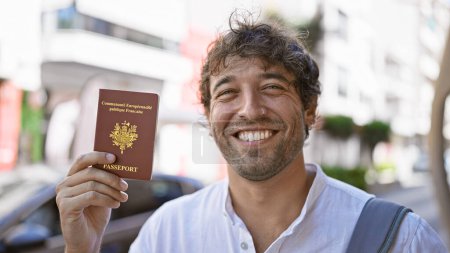 Téléchargez les photos : Beau barbu aux yeux verts tenant un passeport français dans une rue urbaine ensoleillée. - en image libre de droit