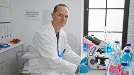 Photo for Happy middle age man, confident hispanic scientist, grinning while analyzing sample through microscope in bustling medical lab - Royalty Free Image