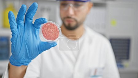 Photo for A young man with a beard examines a petri dish in a laboratory setting, illustrating research or medical analysis. - Royalty Free Image