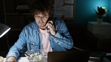Photo for Hispanic man in denim talking on phone at night in office with salad and laptop - Royalty Free Image