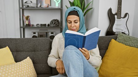 Foto de Una mujer madura en un hiyab absorta en leer un libro mientras está sentada en un sofá en una acogedora sala de estar, evocando una sensación de ocio y tranquilidad. - Imagen libre de derechos
